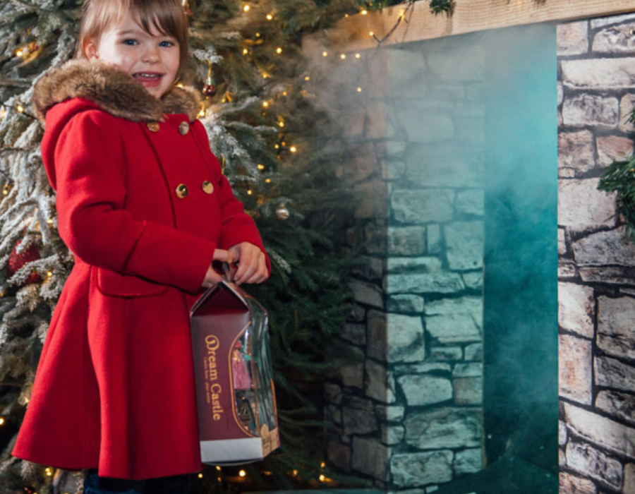 cute little girl with brown hair dressed in a long red coat holding a box with a toy in front of chimney during the best Christmas event in Derby