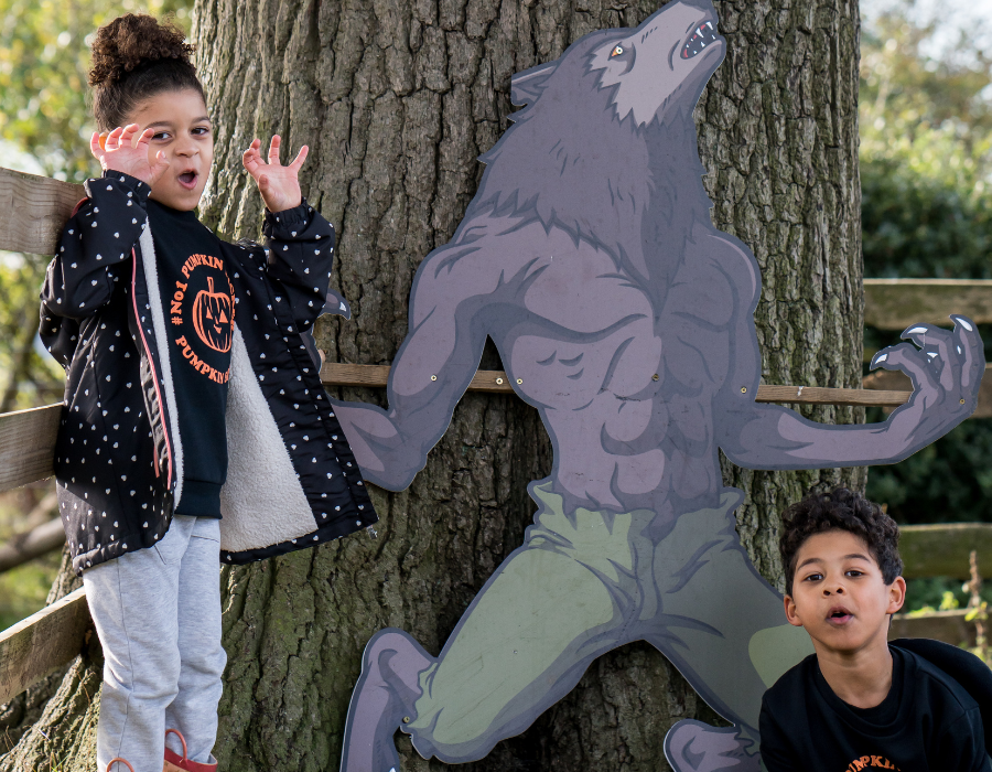 two kids posing for the camera growling to the camera the girl on the left is holding her hands in the air pretending she has claws and a cut-board image of a werewolf stack to the tree in between the kids as part of Alchemist Trail during the best Halloween event in Derby