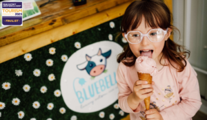 A cute little girl standing in front of ice cream stand with black and white cow logo and wearing pink dress with pink glasses whilst licking pink ice cream