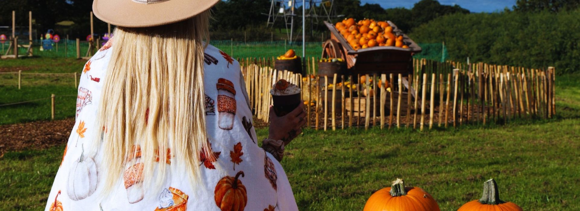 Woman with long blond hair sitting with her back towards camera sipping coffee surrounded by orange pumpkins at farm in Derby